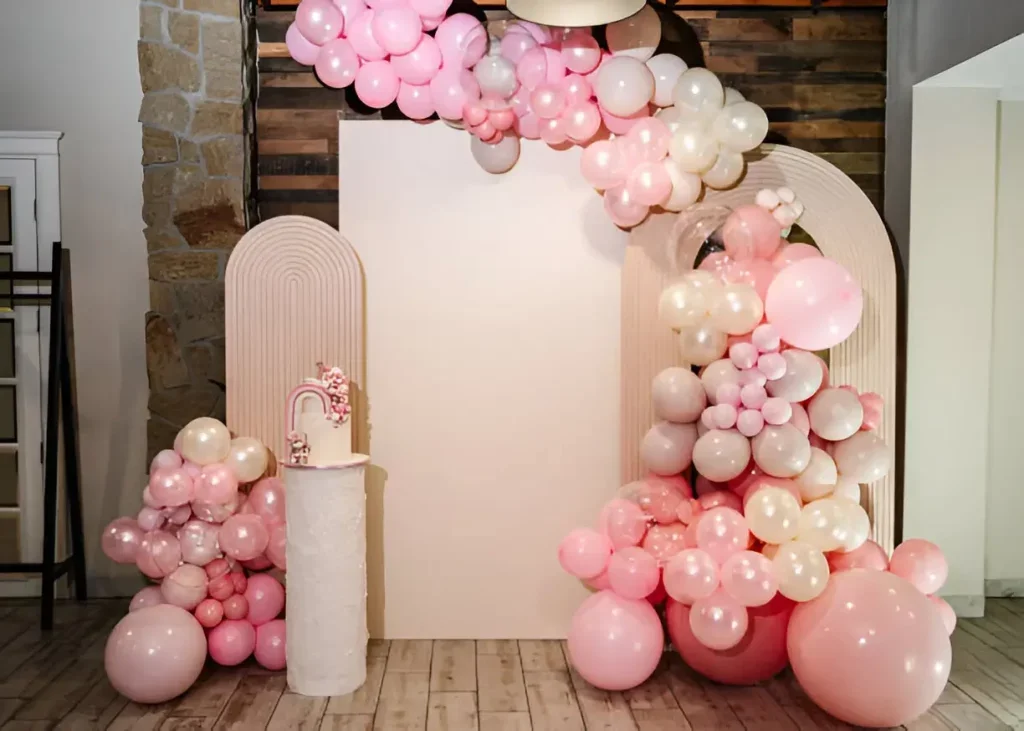 Decorative balloon arch in pink and white with a white backdrop and a small table with a pink rainbow decoration.