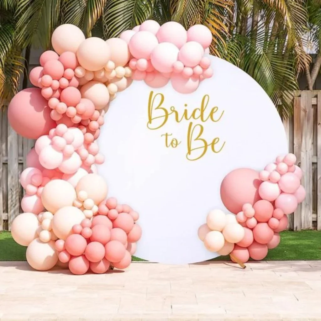 Round white and pink balloon arch surrounding a white circular sign with gold lettering that says "Bride to Be"; outdoor setting with wooden fence and palm trees in the background.