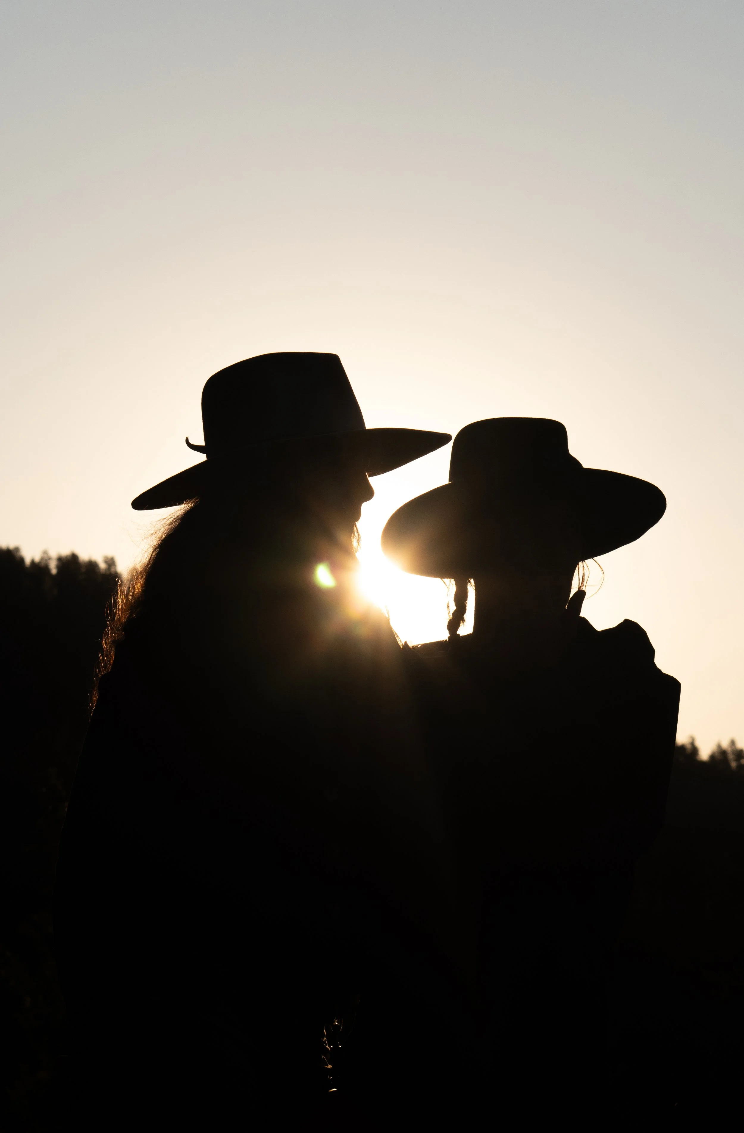 Silhouettes of two women wearing hats, standing close together outdoors at sunset or sunrise.
