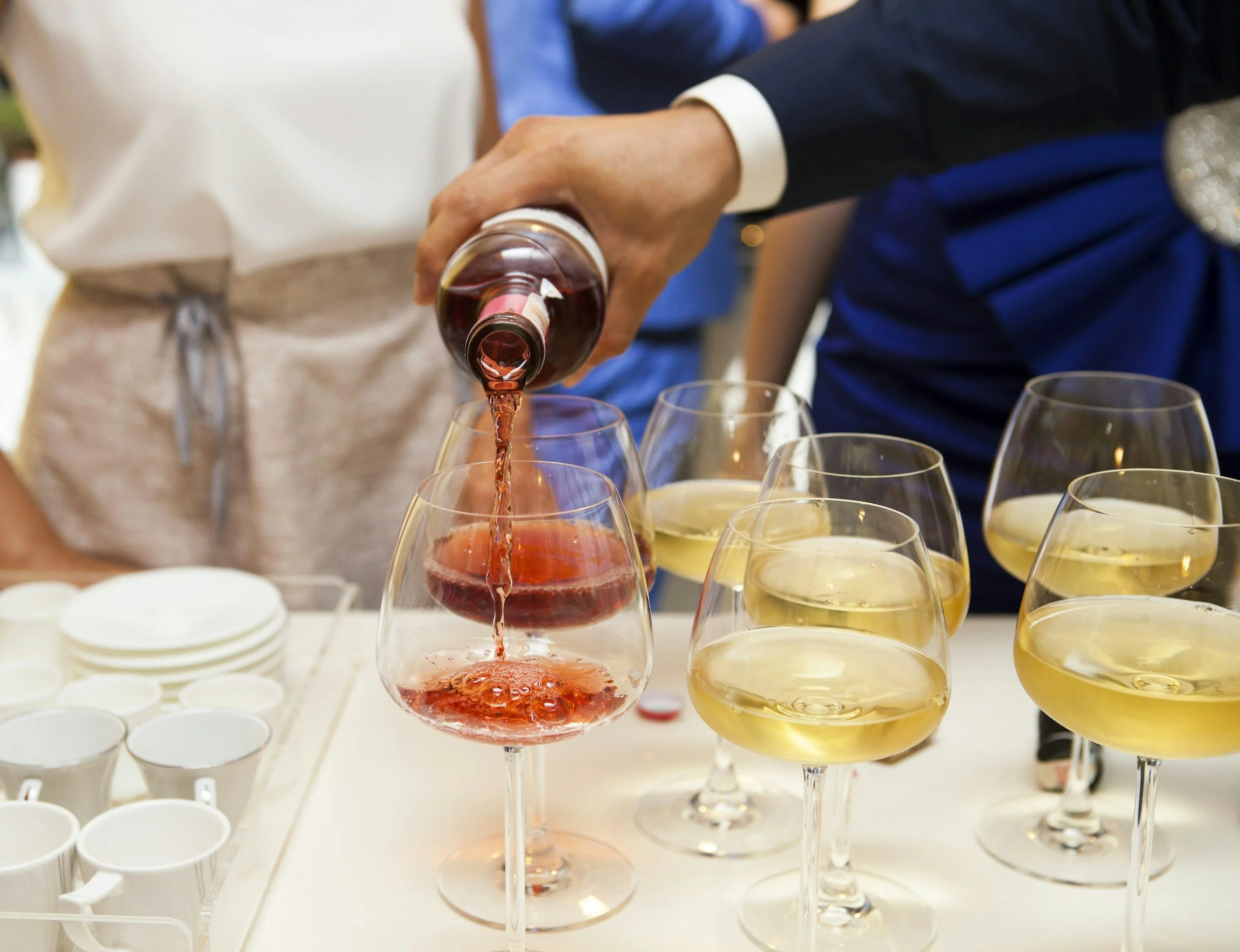 Person pouring red wine into glass at a social gathering with multiple wine glasses on the table.