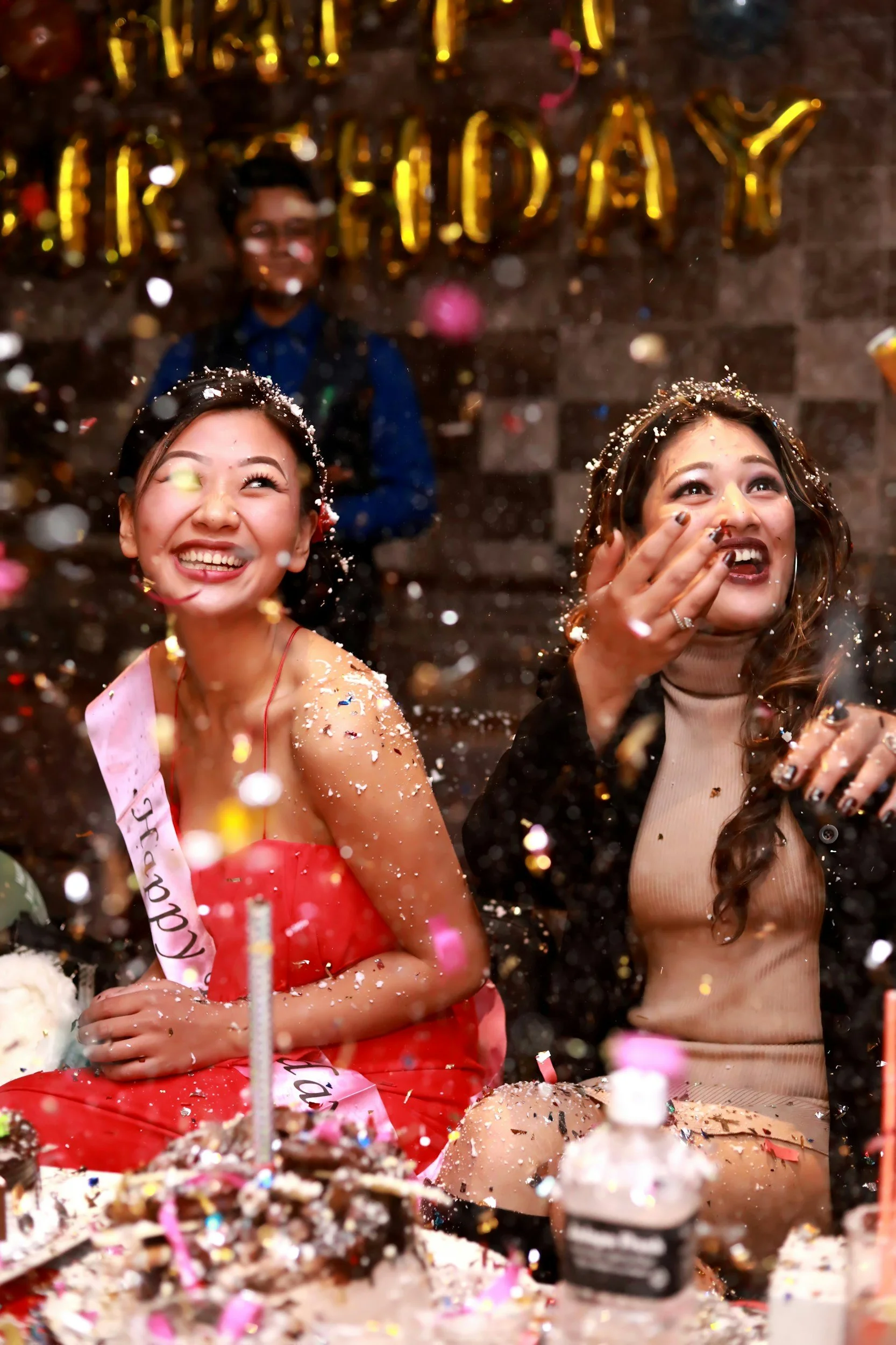 Two women celebrating at a birthday party with confetti, cake, and decorations, one wearing a sash and a red dress, the other in a beige dress with a black jacket.