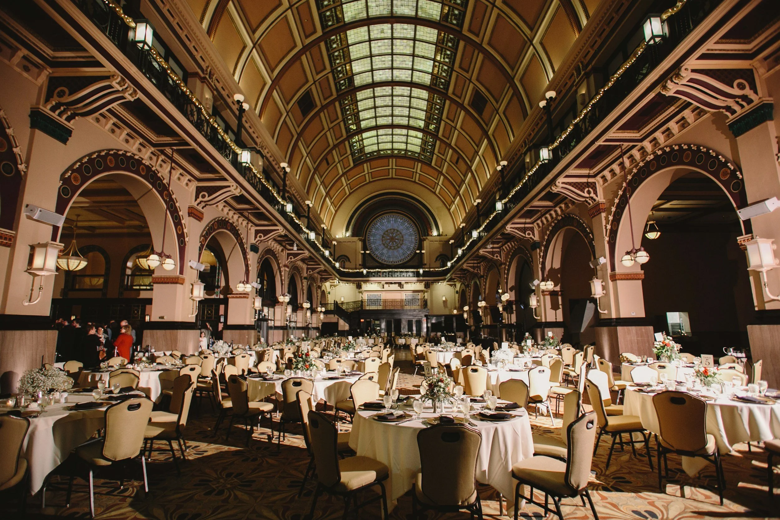 Elegant banquet hall with round tables draped in white cloths, decorated with floral centerpieces, set for a formal event, featuring high arched ceilings, ornate architecture, and large stained glass window at the front.