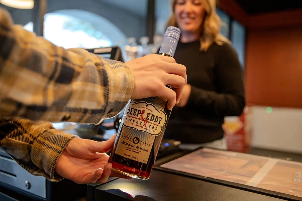Person in a plaid shirt handing a bottle of Deep Eddy Sweet Tea vodka to a smiling woman behind a counter in a restaurant or bar.