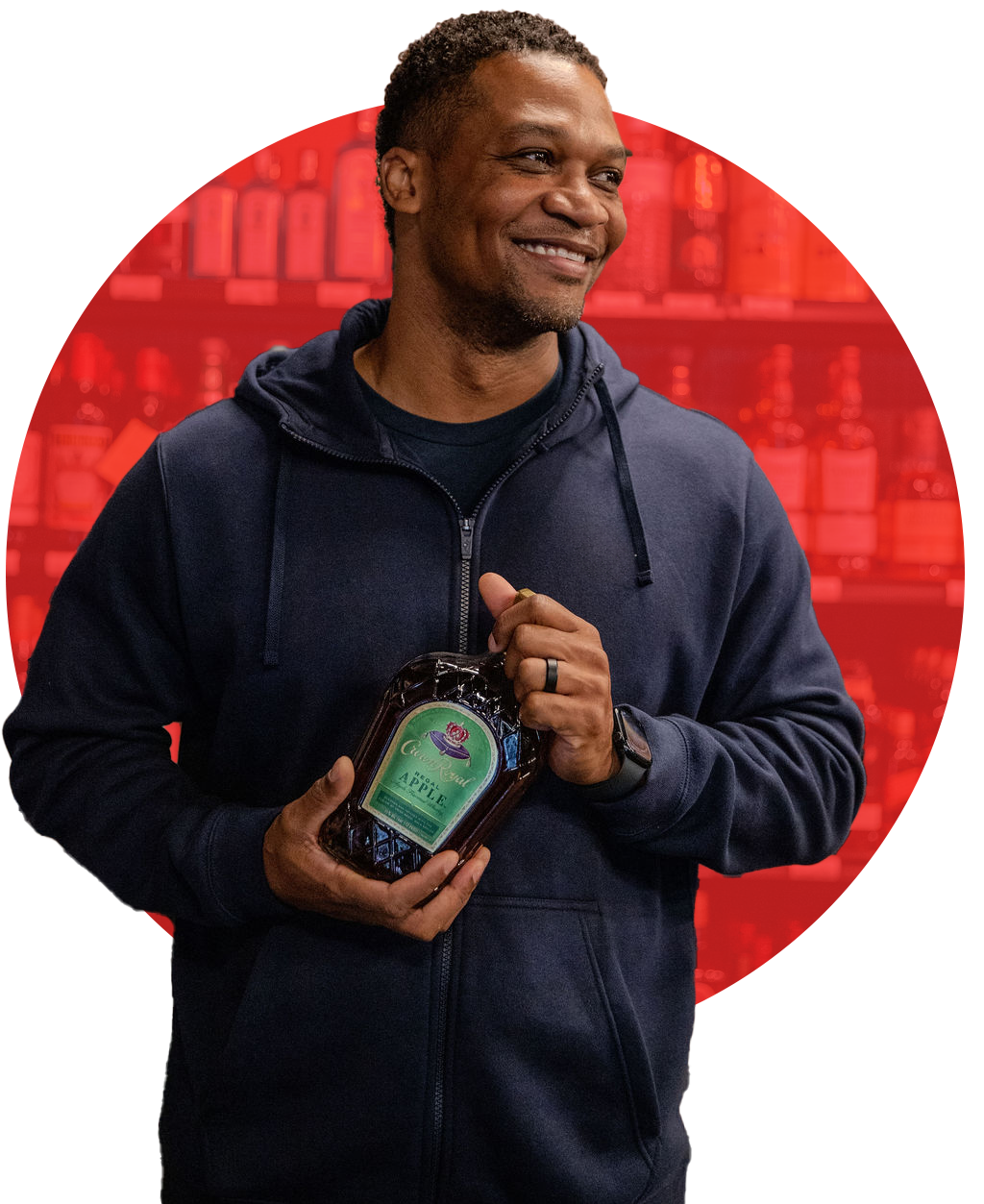 A smiling man in a navy hoodie holding a bottle of apple syrup, standing in front of a red background with shelves of bottles.