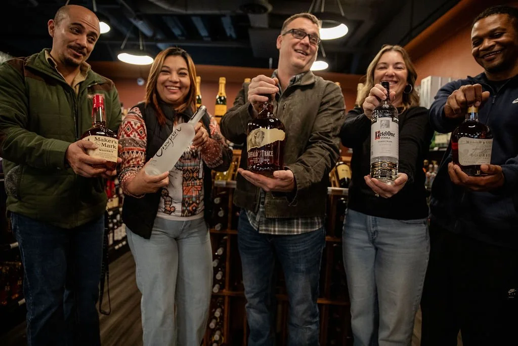 Five people standing in a liquor store holding bottles of alcohol and smiling.