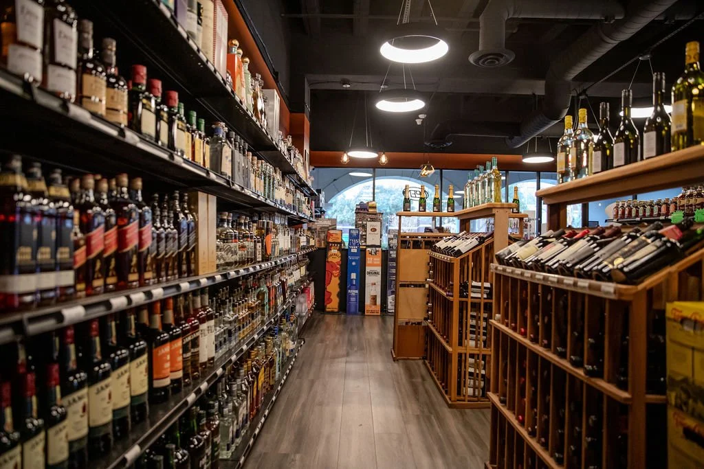 A wine and liquor store aisle with shelves filled with bottles of wine and spirits, wooden wine racks on the right, and a window at the end allowing natural light.