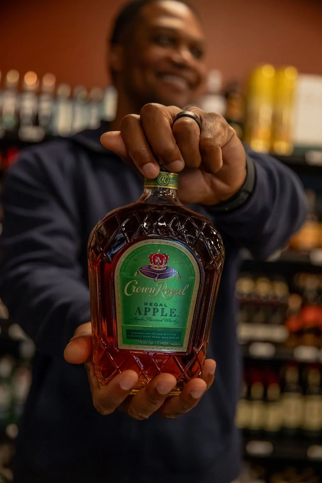 A man holding a Crown Royal Regal Apple whiskey bottle, smiling in a bar or liquor store environment.