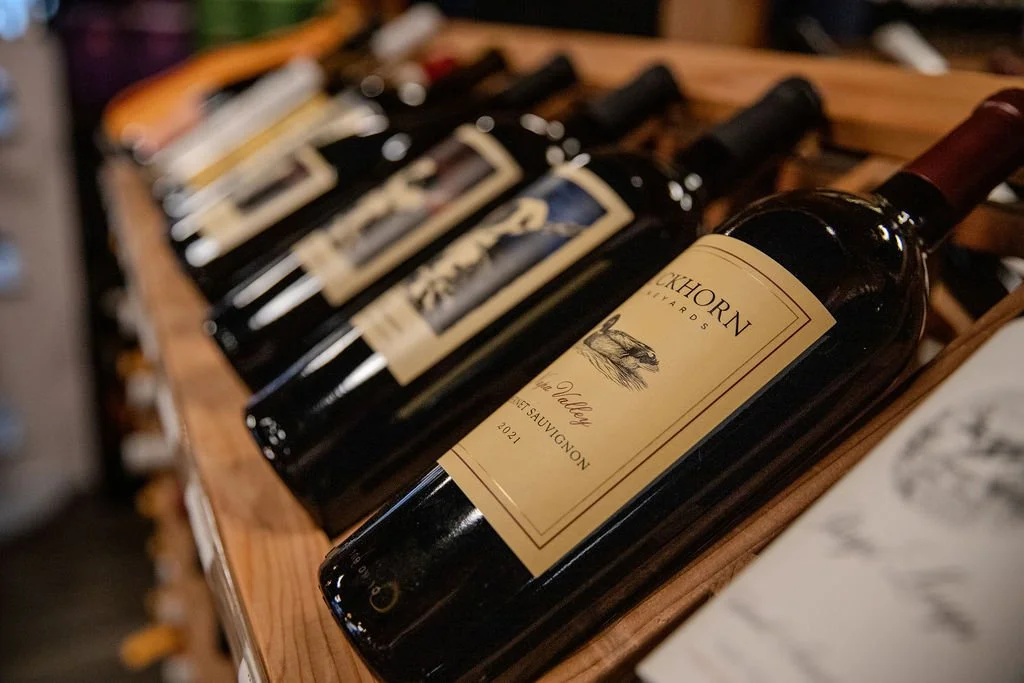 Multiple bottles of wine on a wooden shelf, with the front bottle labeled 'Akhorn Vineyard' and 'Cabernet Sauvignon'.