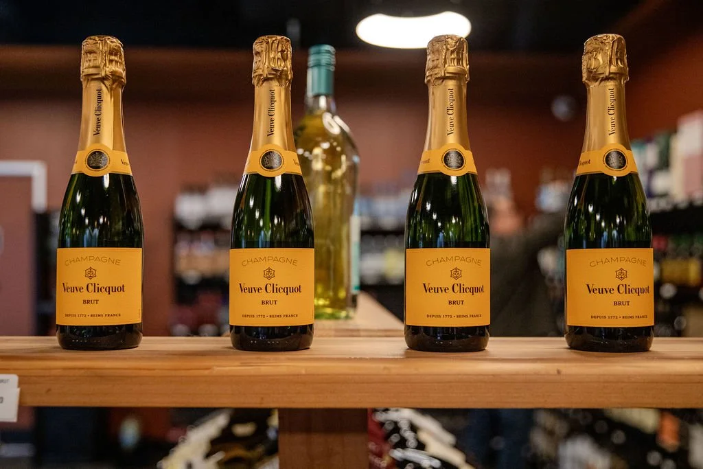 Four bottles of Veuve Clicquot champagne on a wooden shelf in a store, with a blurred background of other bottles and shelves.