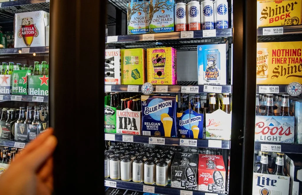 View of a snack and beverage refrigerator in a store aisle with various beer cans and boxes, including Heineken, Blue Moon, Coors Light, and Shiner Bock.