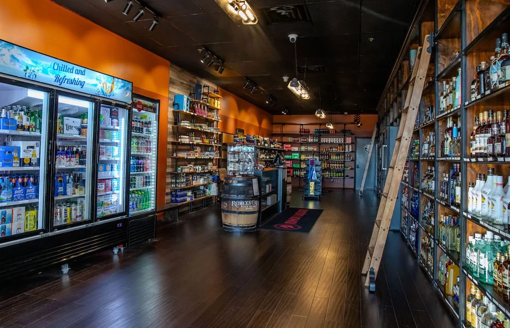 Inside a liquor store with shelves stocked with bottles of alcohol, a refrigerator filled with bottled beverages, and warm lighting.