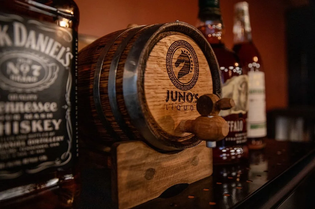 A small wooden barrel labeled 'Juno's Liquor' with a horse's head logo, surrounded by bottles of liquor on a bar counter.