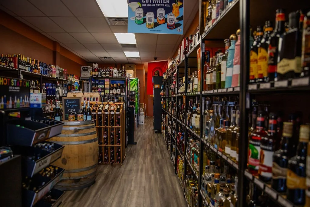 A liquor store aisle with shelves stocked with various bottles of alcohol, a display with wine bottles, and a barrel with beers in the foreground.