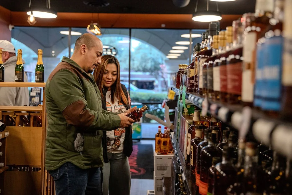 Two people shopping for liquor in a store, looking at a bottle on the shelf.