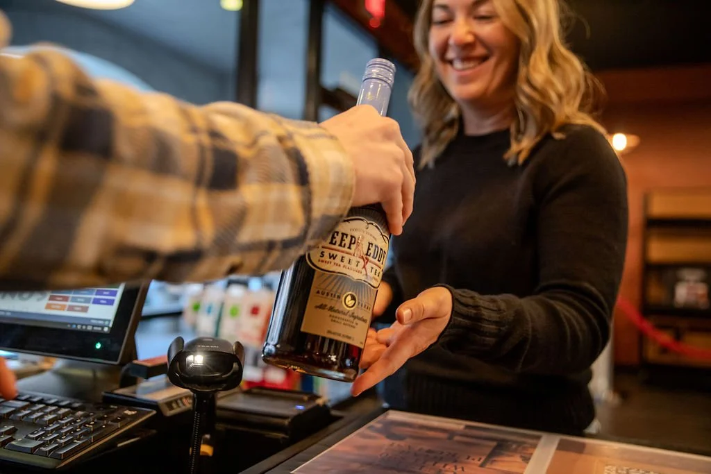 A woman is smiling as she receives a bottle of Deep Eddy sweet tea vodka from a cashier at a retail store or bar counter.