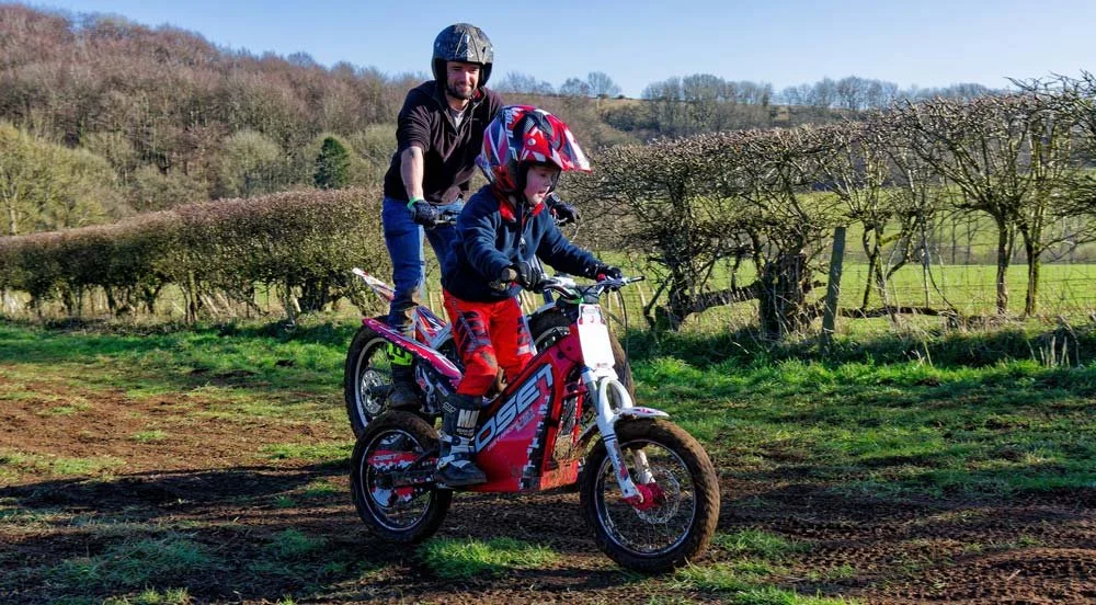 A young rider practising balance on a trials bike with an adult standing beside them on a sunny field.