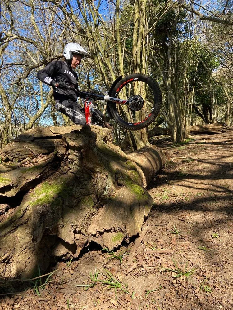 A trials rider climbing over a large fallen tree trunk during practice in the woods.