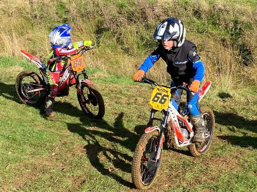 Two young riders practising slow control on a grassy field during a trials session.