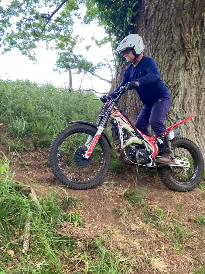 A person wearing a white helmet and blue jacket riding an electric dirt bike on a dirt trail next to a large tree.