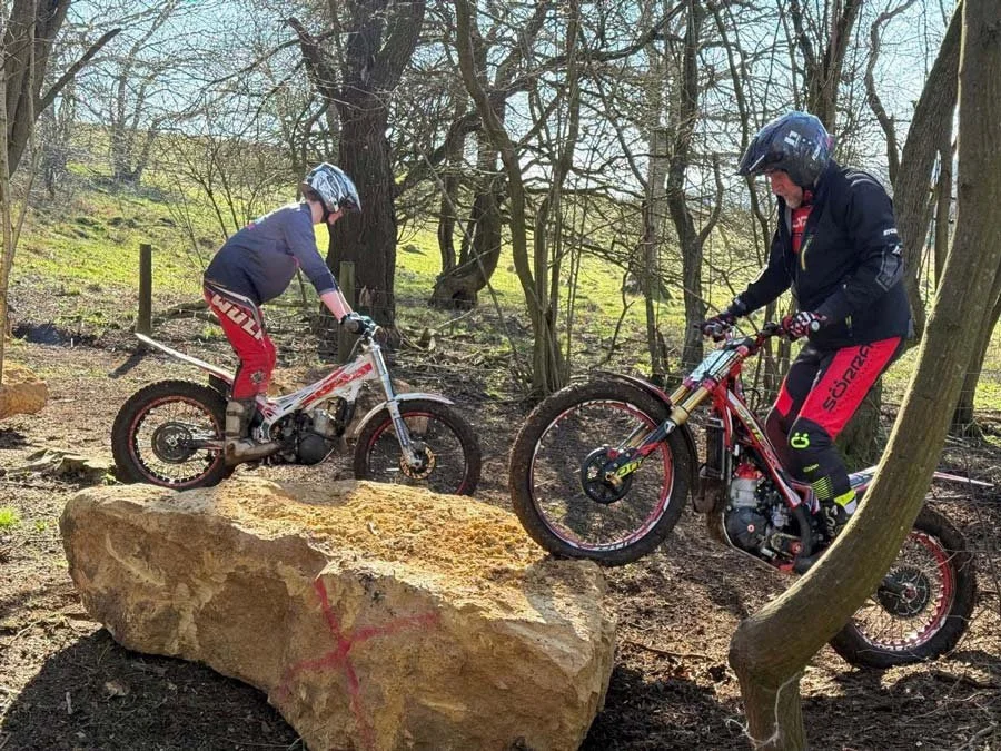 Two people riding trials bikes in a wooded area, one child on a large rock and an adult nearby, both wearing helmets and protective gear.