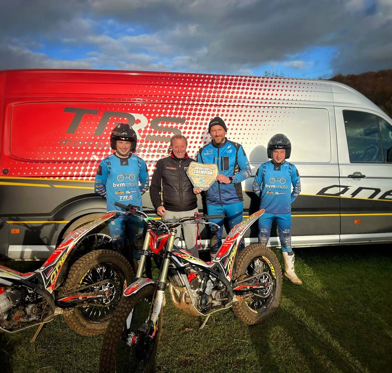 Four trials riders in blue BVM Moto kit stand in front of a TRS team van, with two trials bikes in the foreground. One rider holds a British Champion award plate while the group poses and smiles in the sunlight.