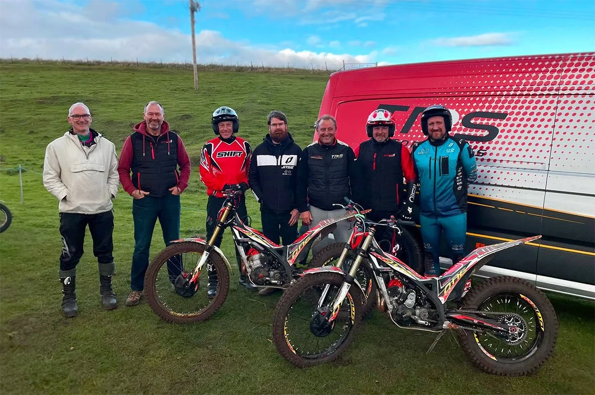 Group of MTC riders and team members standing together beside two trials bikes and a TRS support van on a grassy hill.