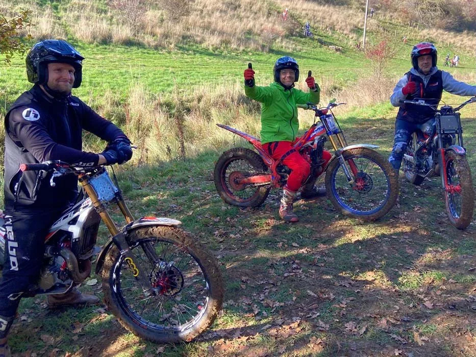Three men in riding gear with helmets standing with dirt bikes on a grassy trail. One man in the middle is giving two thumbs up and smiling, wearing a green jacket and red pants. The other two are on either side, each holding a bike and giving a thum