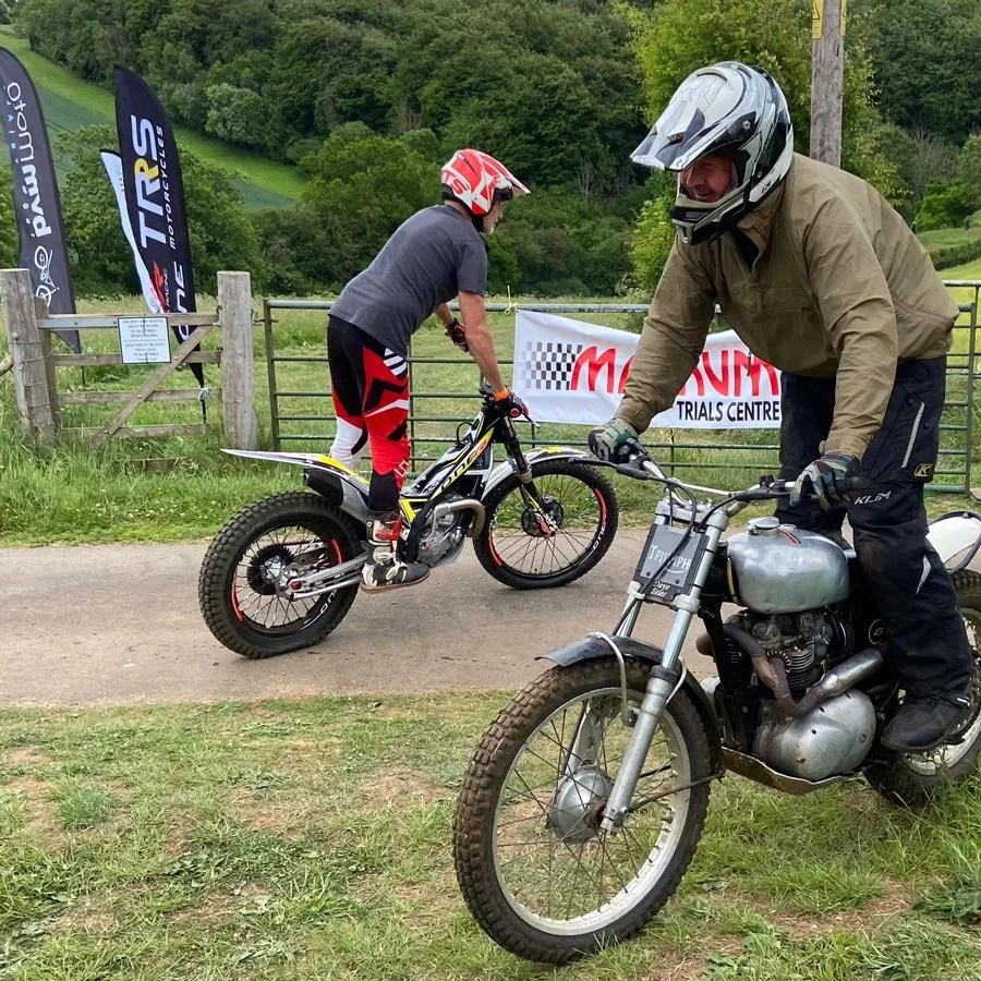 Two trials riders pass each other near the entrance to the venue, one on a modern trials bike and the other on a classic Triumph, with MTC banners and green hills in the background.