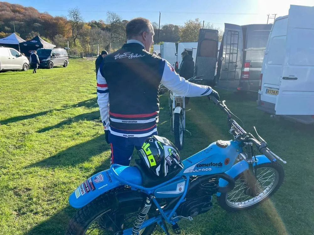 A man in a racing jacket with signatures stands next to a blue vintage motorcycle with a helmet on it, at an outdoor event with several vans and cars in the background.