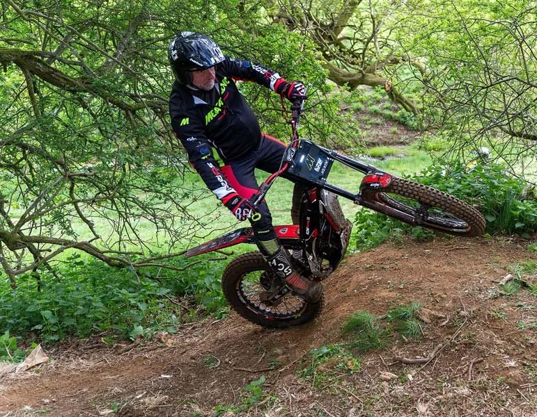 A biker lifting a wheel whilst riding on a dirt trail surrounded by green foliage.