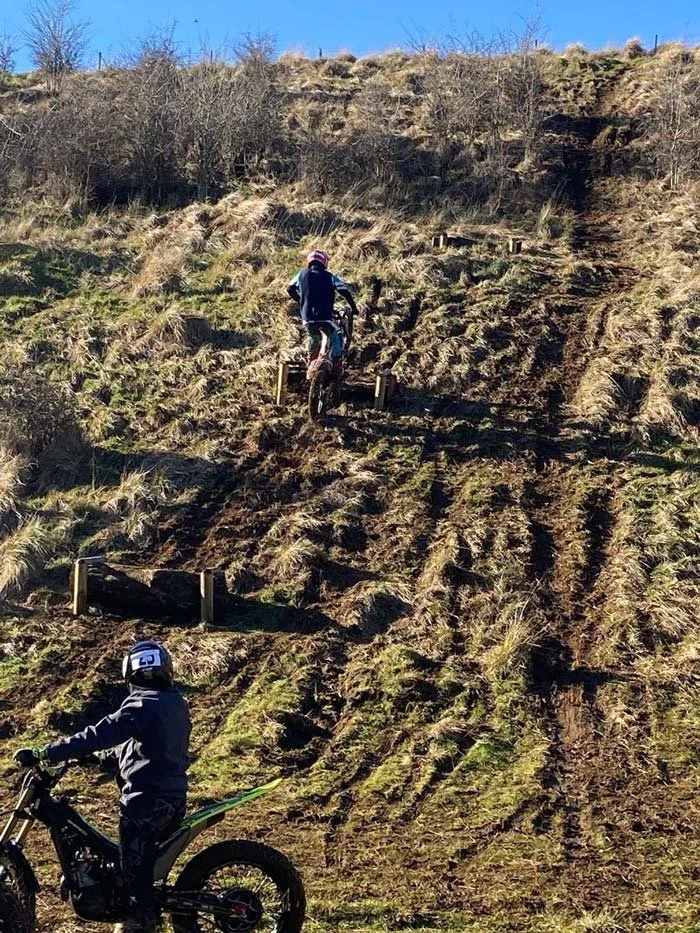 People riding dirt bikes on a steep, muddy hillside with tire tracks and patches of grass.