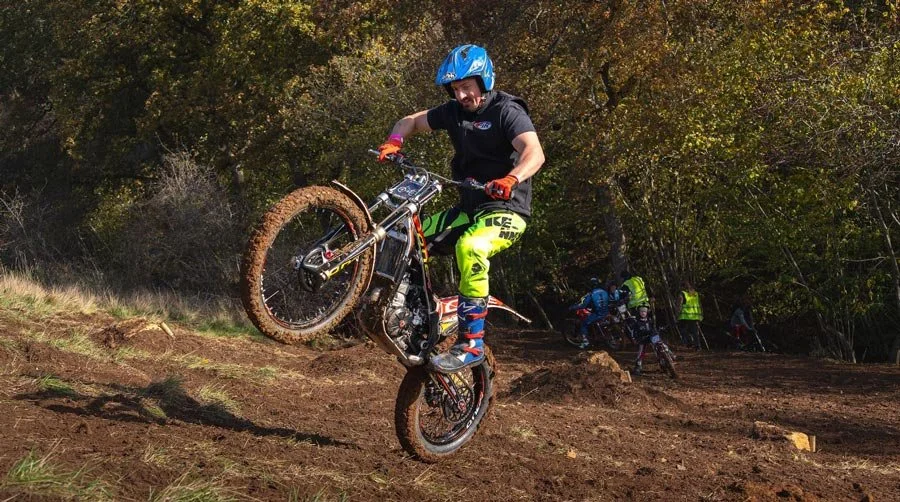 A trials rider lifting the front wheel while tackling a muddy uphill section during practice at Magnum Trials Centre.