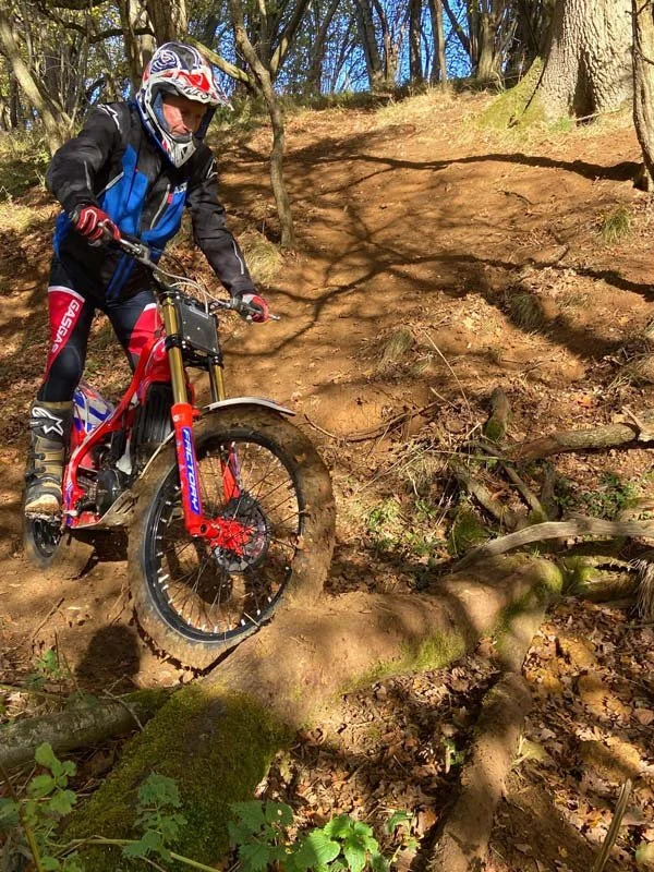 Motorcyclist wearing helmet and riding gear navigating rocky trail in wooded area.