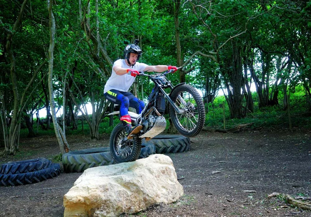 A trials rider climbing onto a large rock with good front wheel lift in a wooded practice area.