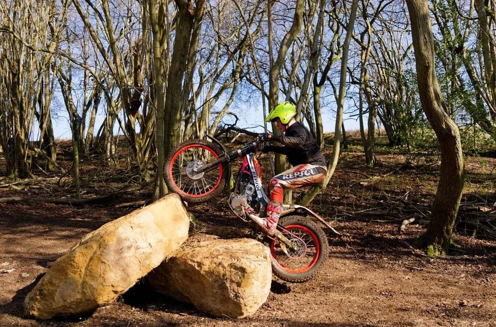 A young trials rider tackling a rock obstacle in a woodland practice zone.