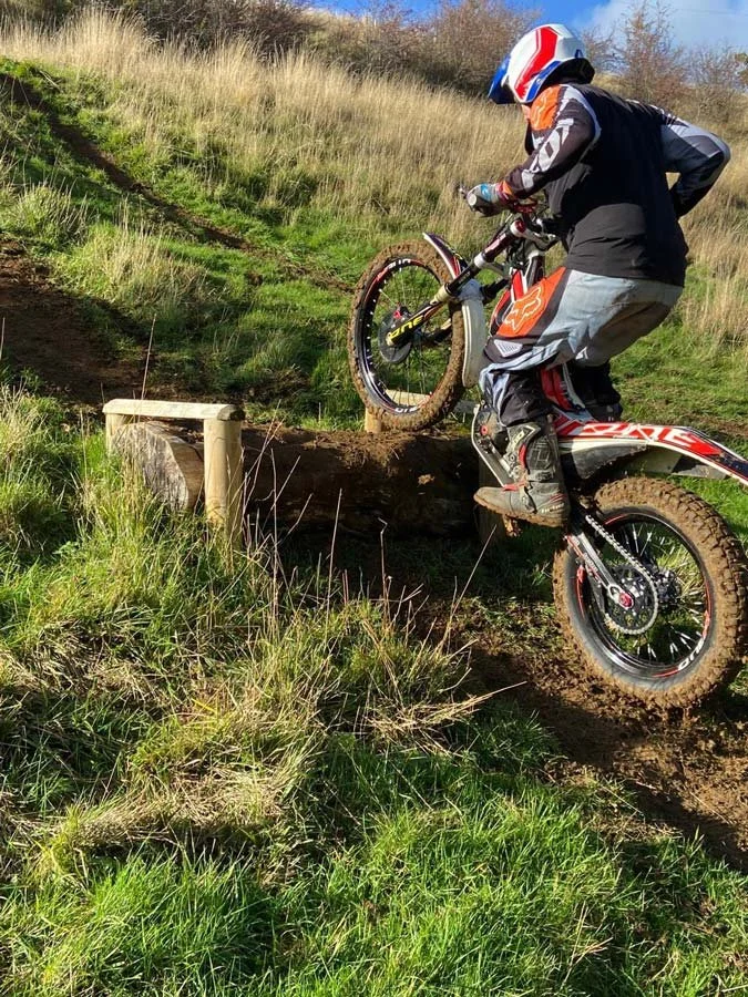 A rider tackling a narrow raised log obstacle, lifting the bike’s front wheel to mount it.