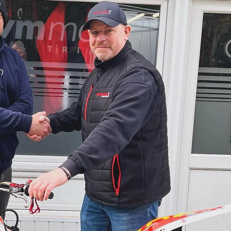 Simon Etheridge in a Magnum Trials Centre black jacket and cap shaking hands with another person, standing next to a bicycle, outside BVM Moto Stroud.