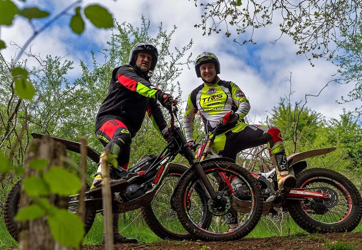 Two trials riders (James Saunders & Heath Brindley) pause together on their bikes in a wooded section of the loop, wearing bright riding gear and helmets, smiling against a backdrop of trees and blue sky.