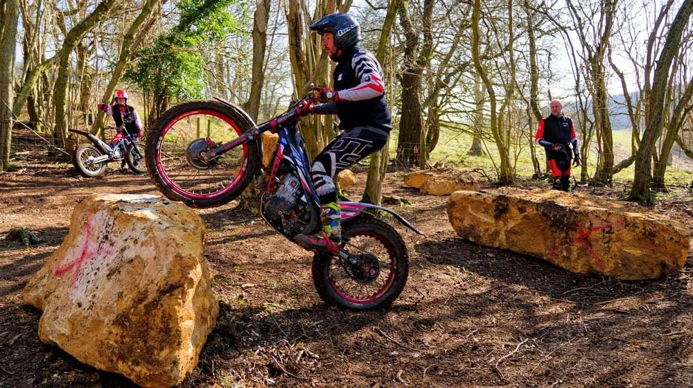 A trials rider performing a controlled rear-wheel lift over rocks in a wooded training area.
