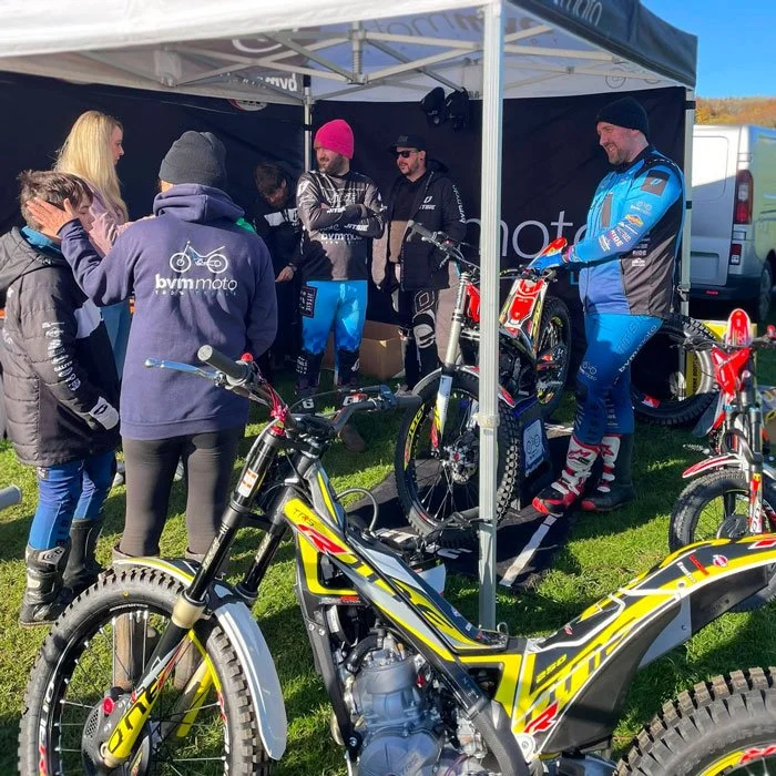 A group of people, including a motocross rider in blue gear, gathered around a dirt bike under a tent at an outdoor event on a sunny day.