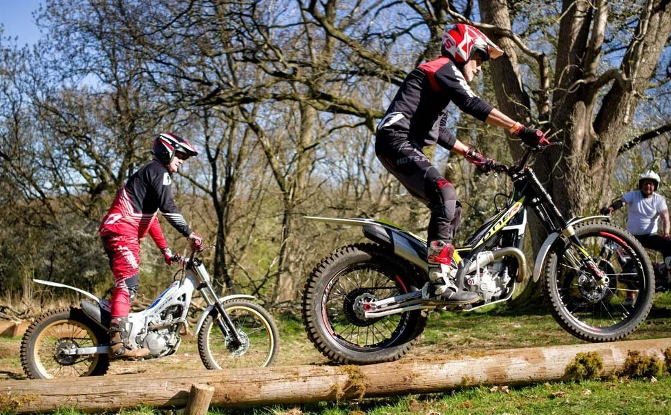 Two experienced trials riders practising balance and control on logs at Magnum Trials Centre, with one rider lifting the bike’s front wheel while another follows on a wooded training section.