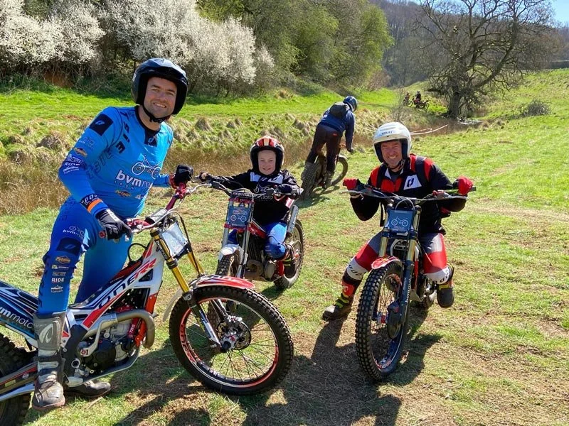 Three riders in biking gear and helmets posing with their bikes on a grassy trail, with two other bikers in the background riding downhill on the trail and trees in a sunny outdoor setting.