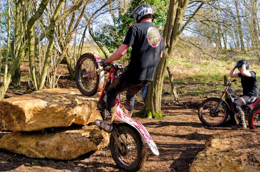 A trials rider climbing onto a rock feature in a wooded practice section at Magnum Trials Centre.