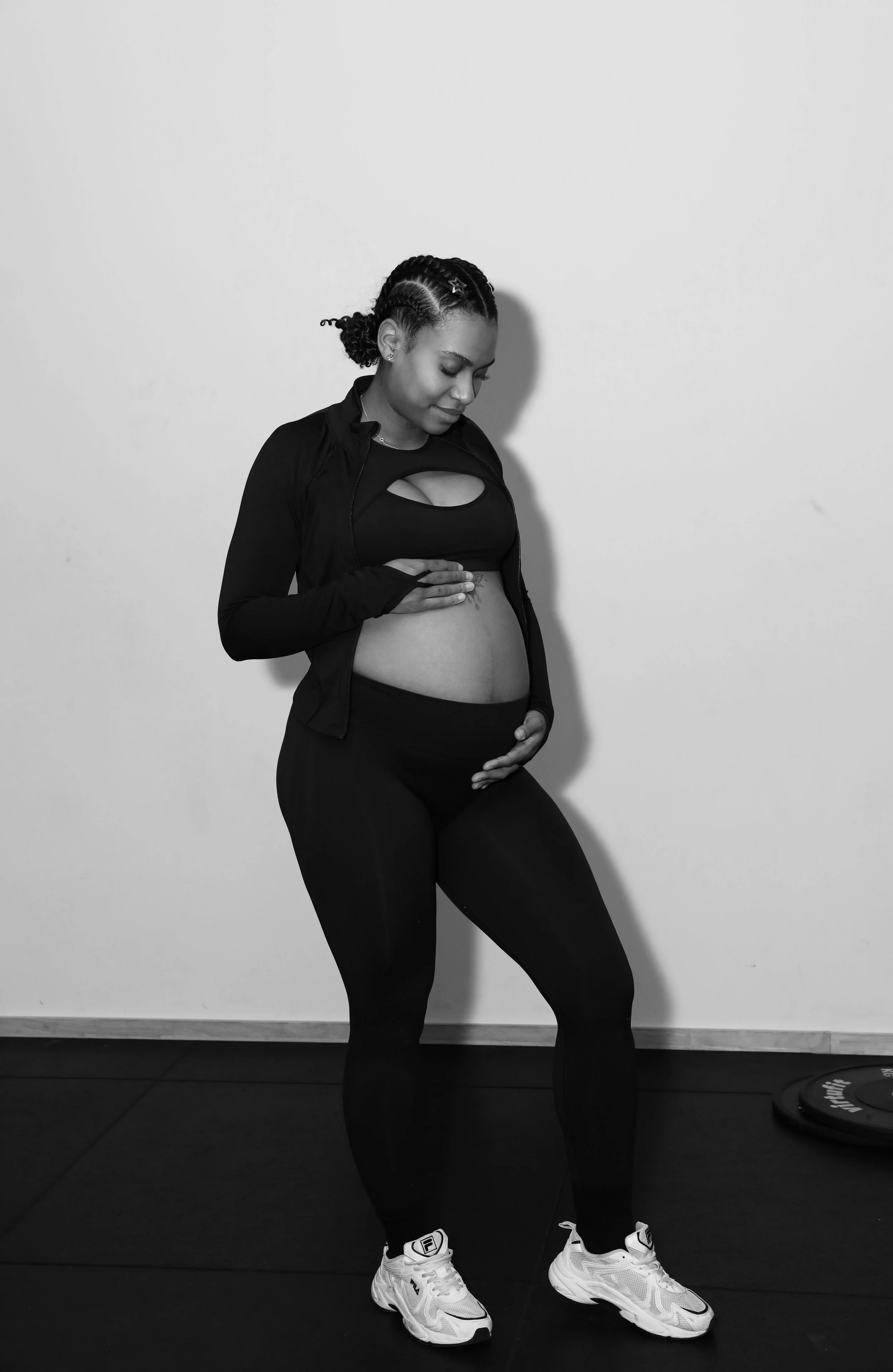 Une femme enceinte en vêtements de sport, les mains sur son ventre, souriant doucement, dans une pièce neutre.