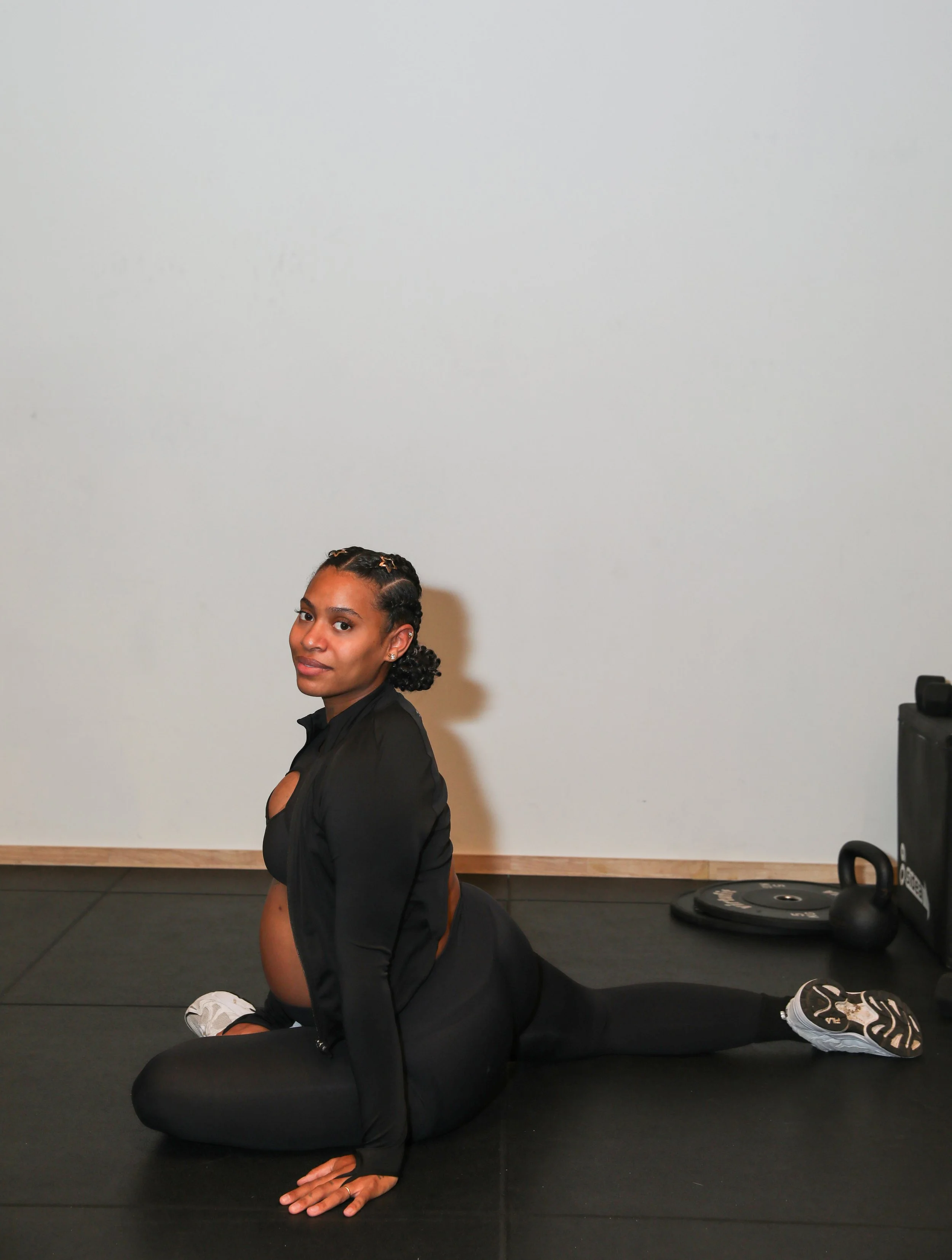 Femme faisant un étirement au sol dans une salle de sport, portant une tenue de sport noire, cheveux courts tressés, avec du matériel de gym en arrière-plan.