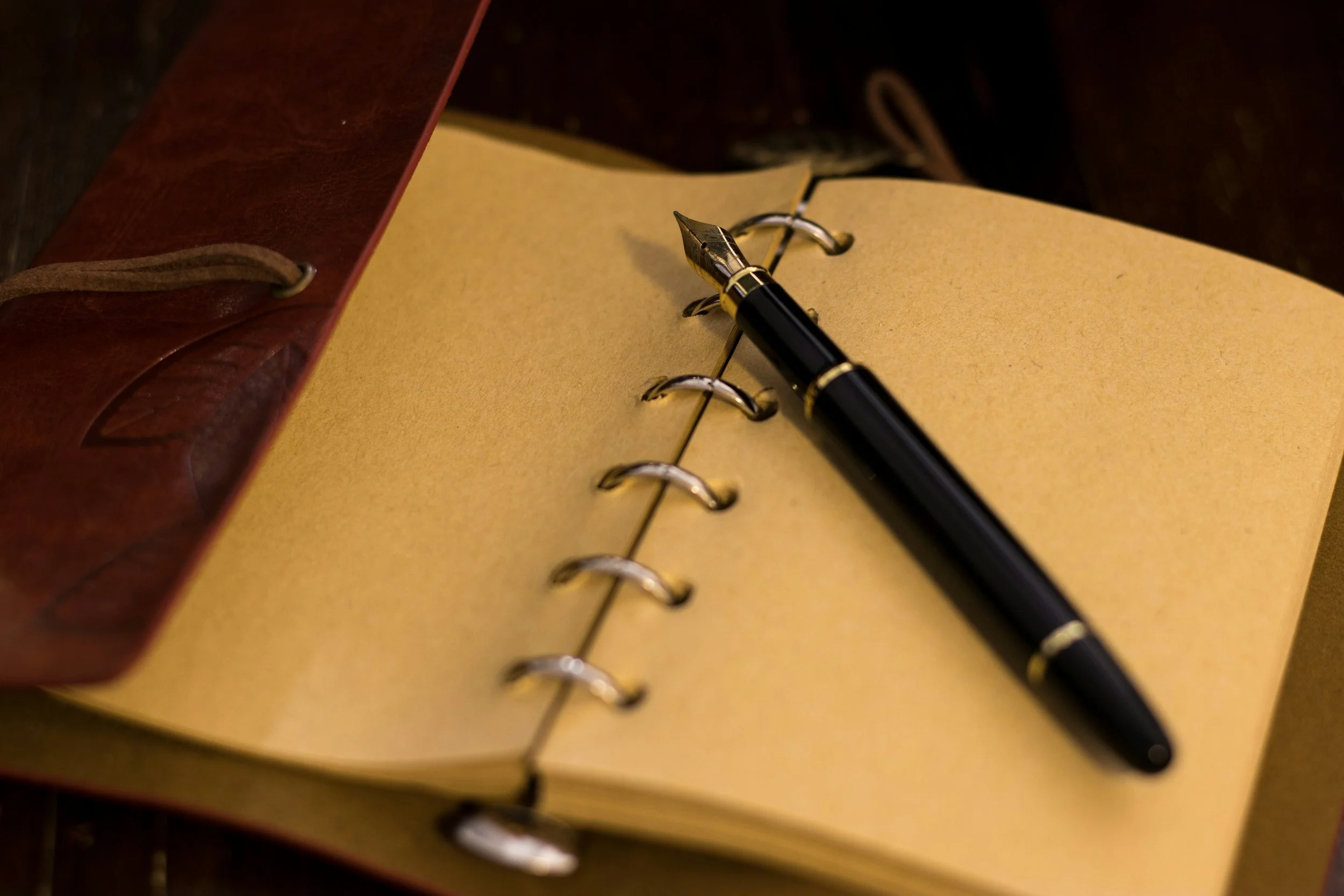 Open leather-bound notebook with yellow pages, a black fountain pen resting on it, and metal rings binding the pages.