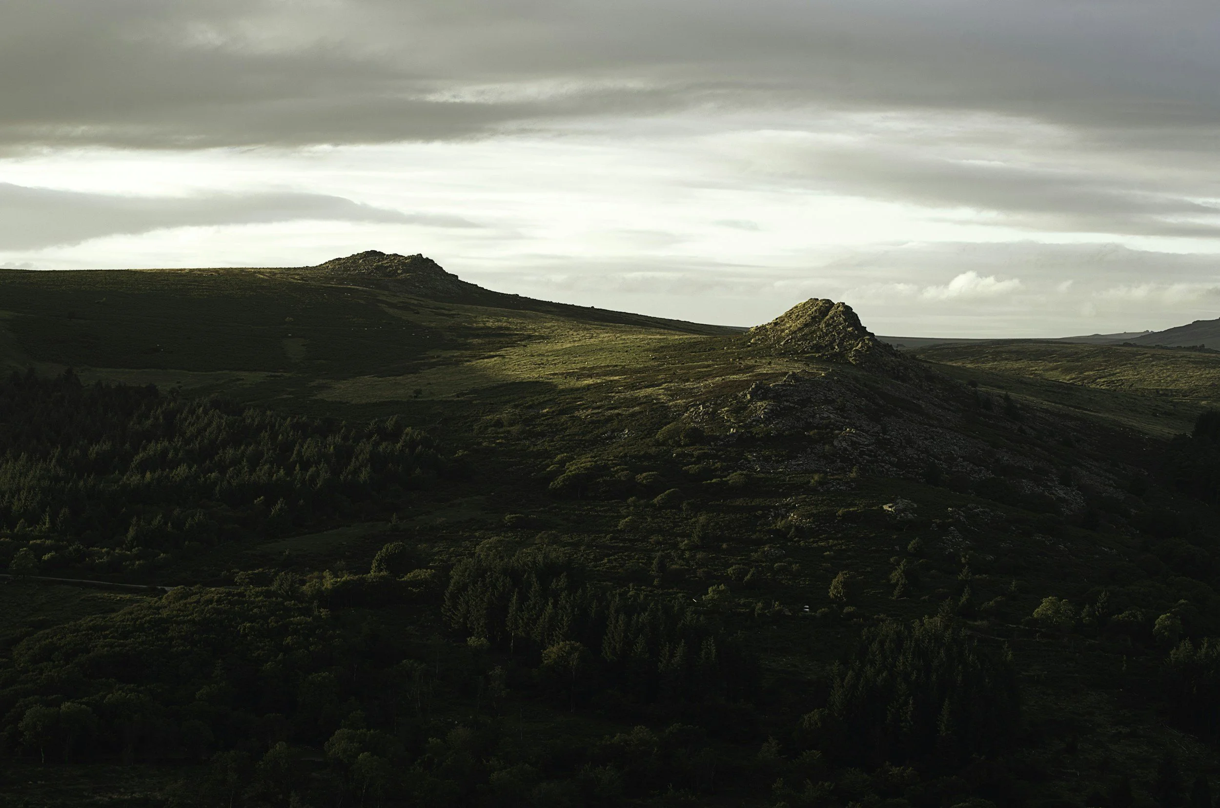 A landscape with rolling green hills and rocky outcroppings under a cloudy sky, with sunlight illuminating parts of the terrain.