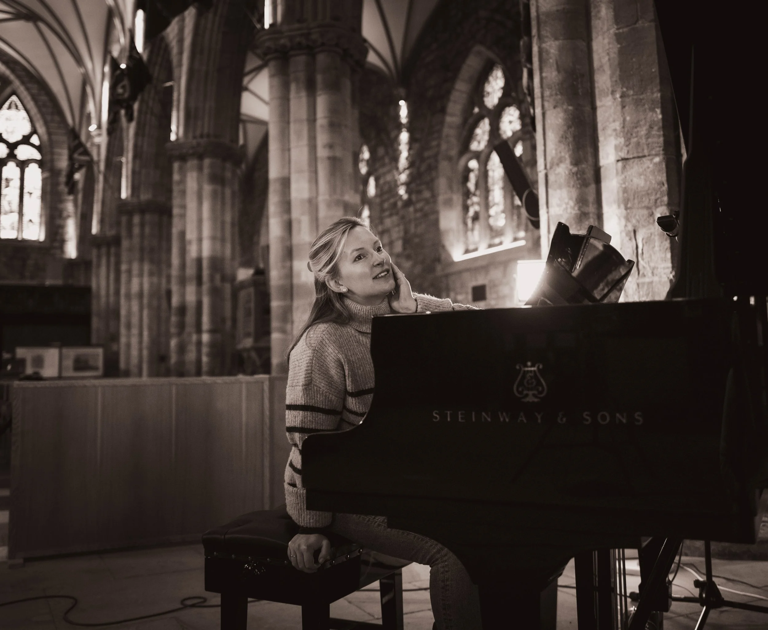 Natalie Burch sitting at a Steinway & Sons grand piano inside a church, with high arched windows and stone columns in the background, appears to be singing or performing.
