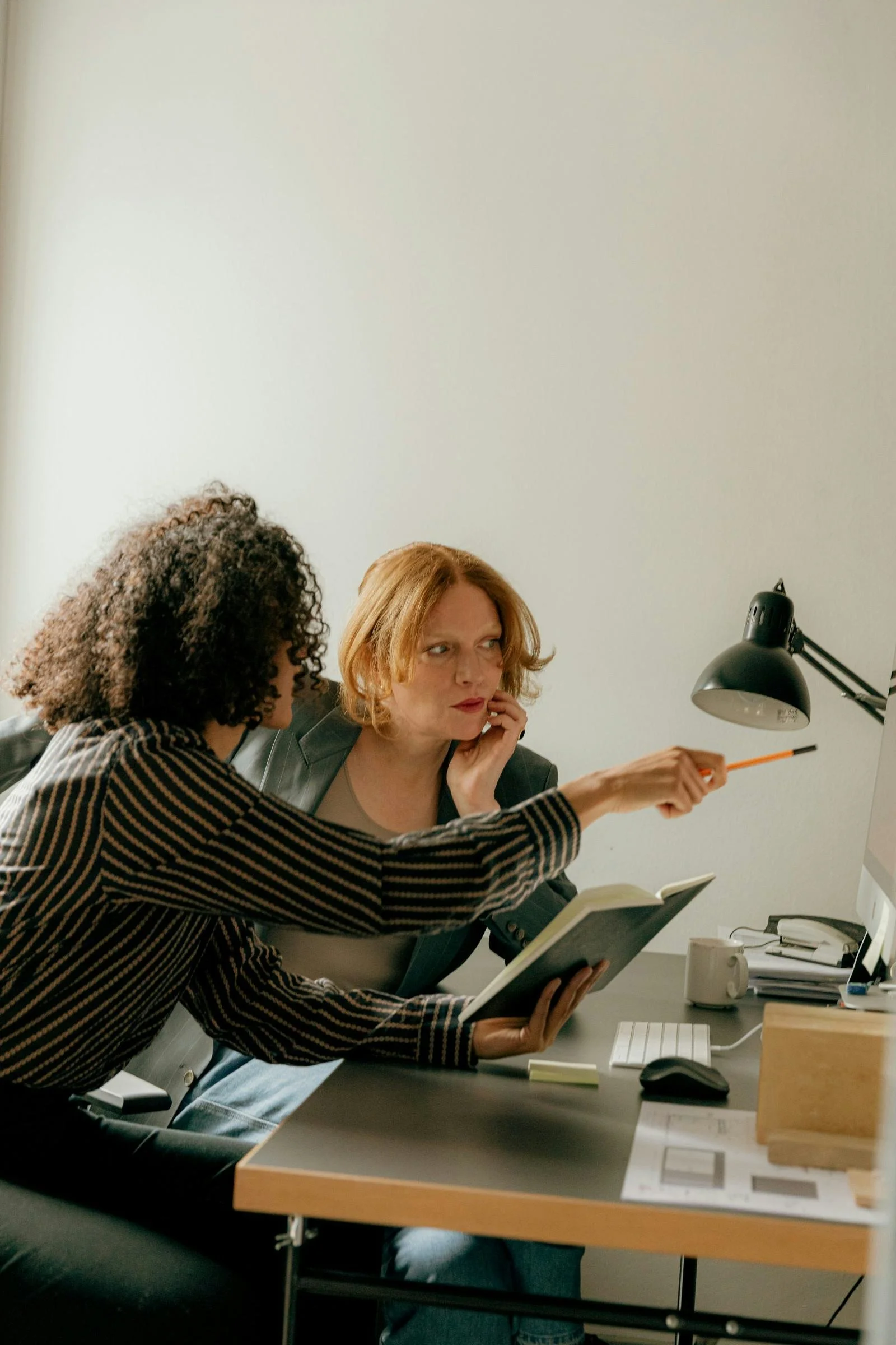 Two women working at a desk with a computer, one pointing at the screen and the other reading a notebook.