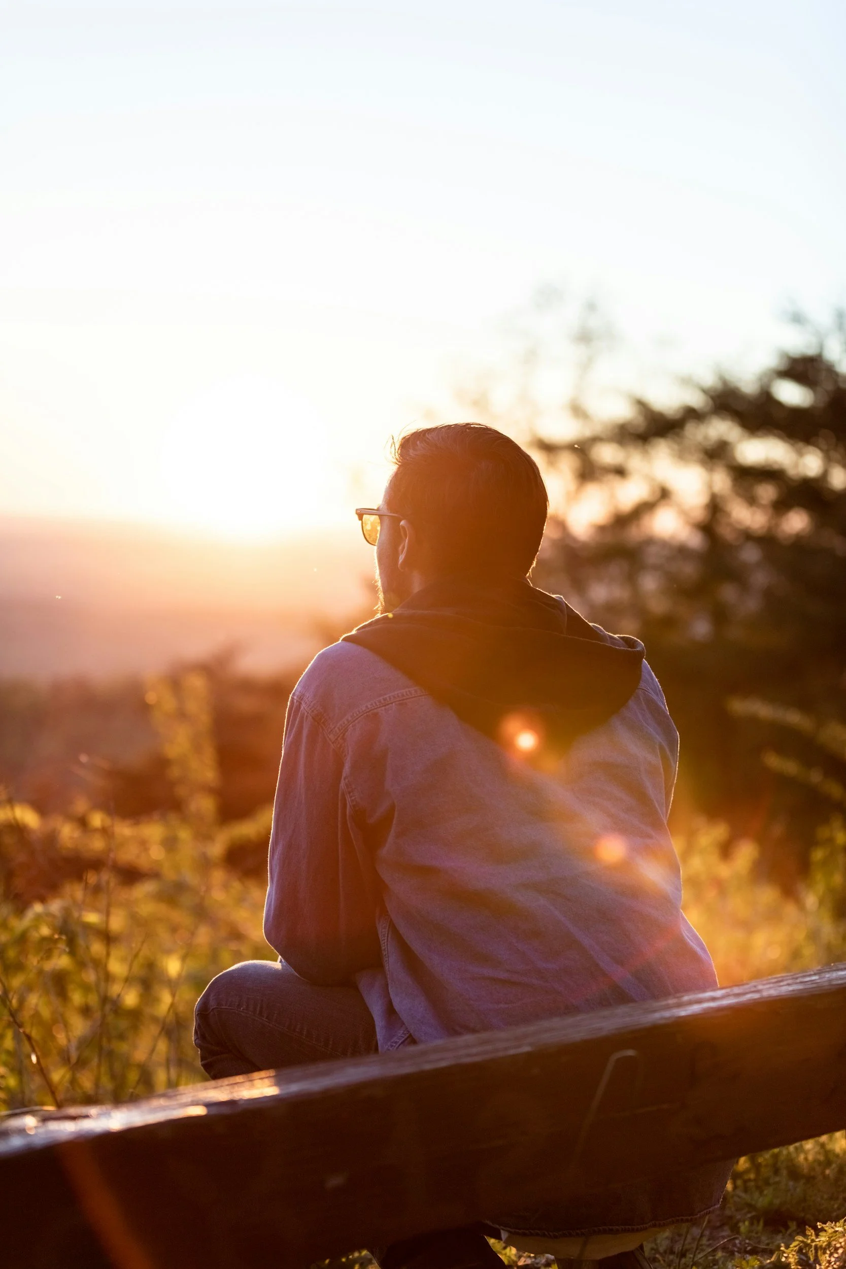 A man sitting on a bench outdoors during sunset, wearing sunglasses and a denim jacket with a hoodie, looking at the sunset.